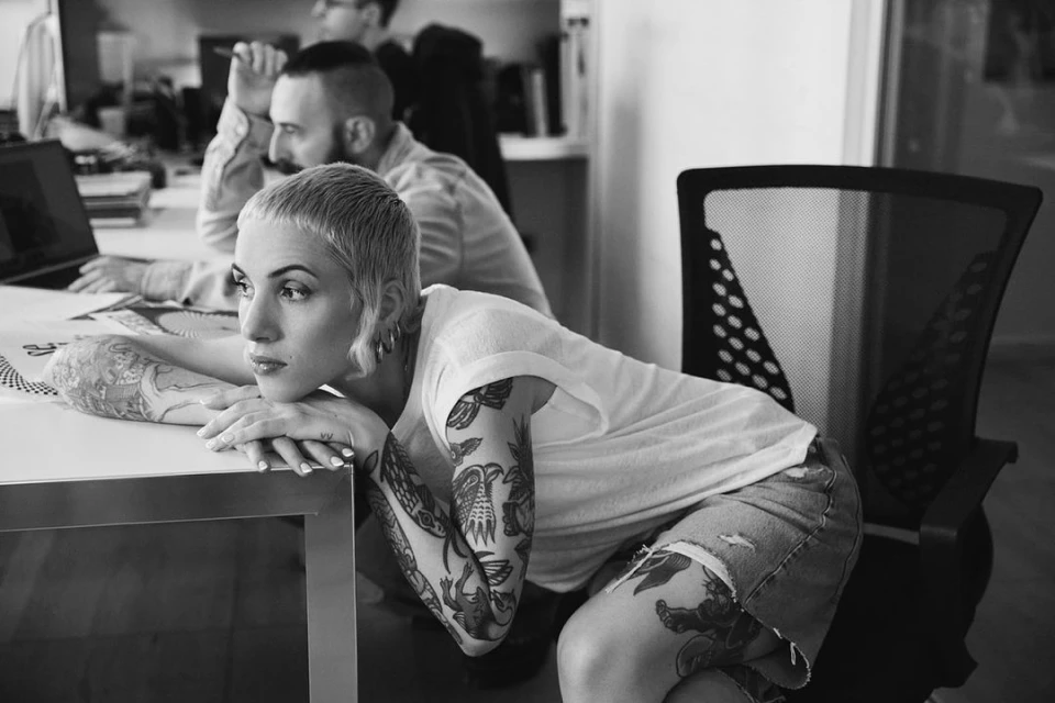 Black and white portrait of a woman with short hair and arm tattoos, leaning on a white desk in an office setting with colleagues.
