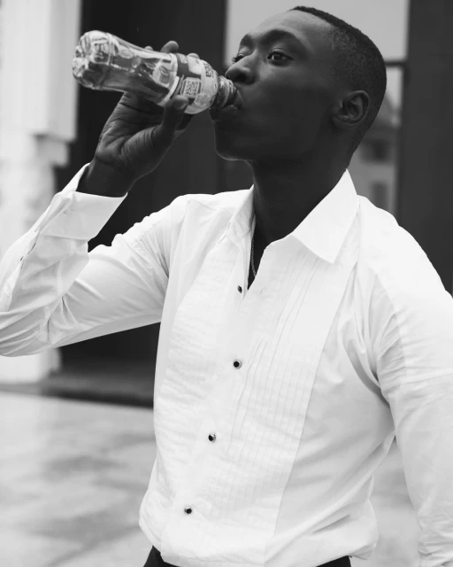 Monochrome portrait of a black man drinking from a plastic bottle outdoors, wearing a white button-down shirt with black buttons.