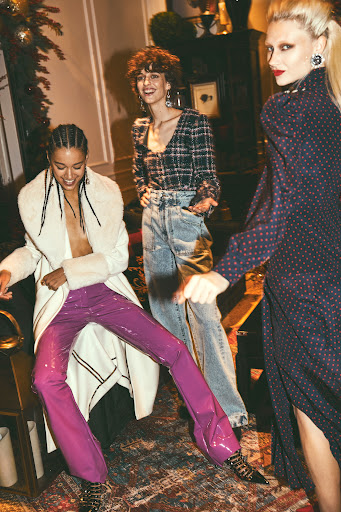 Three fashion models in an indoor setting. One model wears purple pants, one wears jeans, and the third wears a long dress.