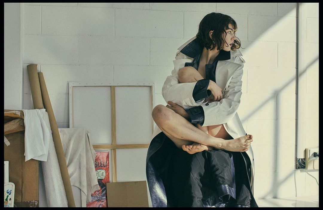 Woman in a coat seated on a black fabric, posing in an artist's studio with painting supplies, casting shadows with natural light.