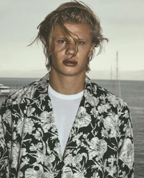 Close-up portrait of a blonde man wearing a black and white floral patterned blazer and white t-shirt against a coastal water background