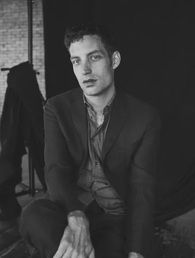 Monochrome portrait of a man in a dark suit jacket and button-down shirt seated in a studio setting