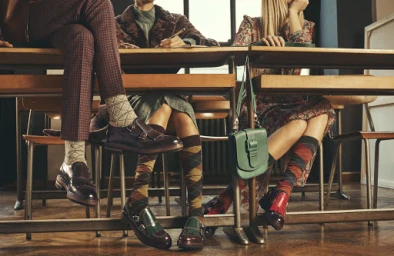 Three models seated at wooden desks wearing patterned socks and leather shoes, photographed by Stefano Galuzzi.