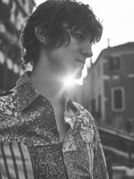 Monochrome portrait of a young man wearing a patterned patchwork button-down shirt outdoors with sunlight glowing behind him.