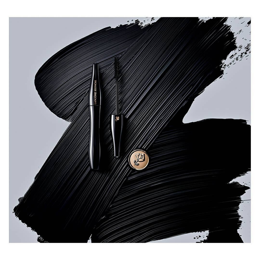 Lancôme mascara tube and applicator brush sit on a dark textured smear against a light gray backdrop in this studio product shot.