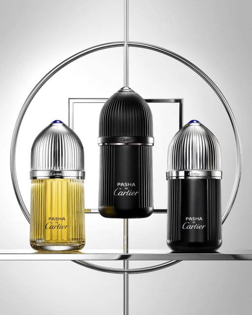 Three Pasha de Cartier perfume bottles in black and gold, displayed on a tiered chrome stand against a graduated gray background.