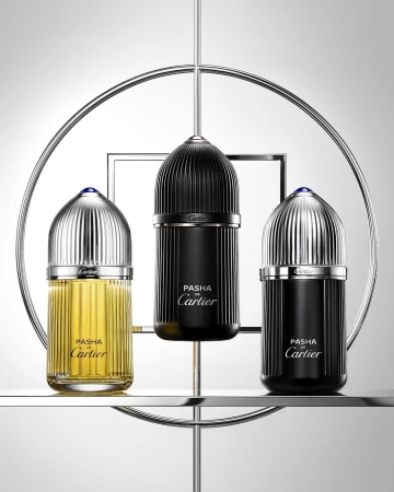 Three Pasha de Cartier perfume bottles in black and gold, displayed on a tiered chrome stand against a graduated gray background.