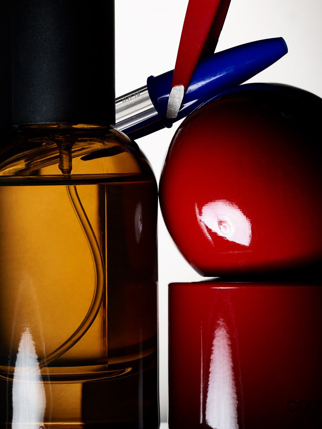 Still life featuring an amber glass bottle, red sphere and cylinder, and a blue and silver pen against a white background in a studio.