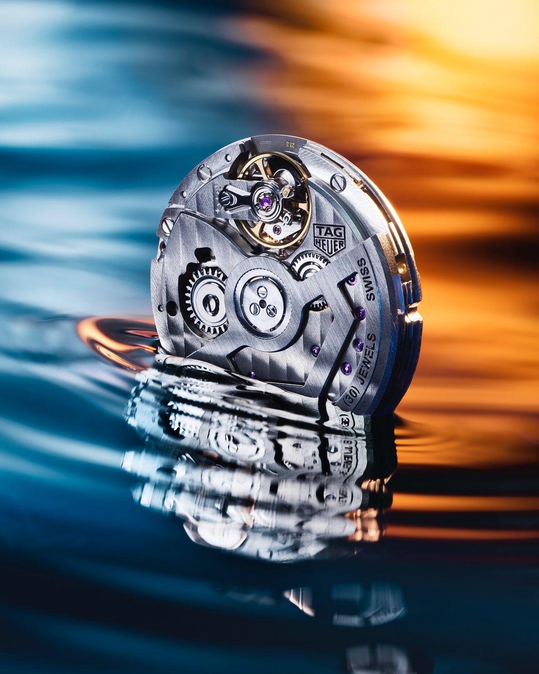 Close-up of an intricate watch movement mechanism, gears visible, and positioned above the reflective water surface. Watch face has a Tag Heuer logo.