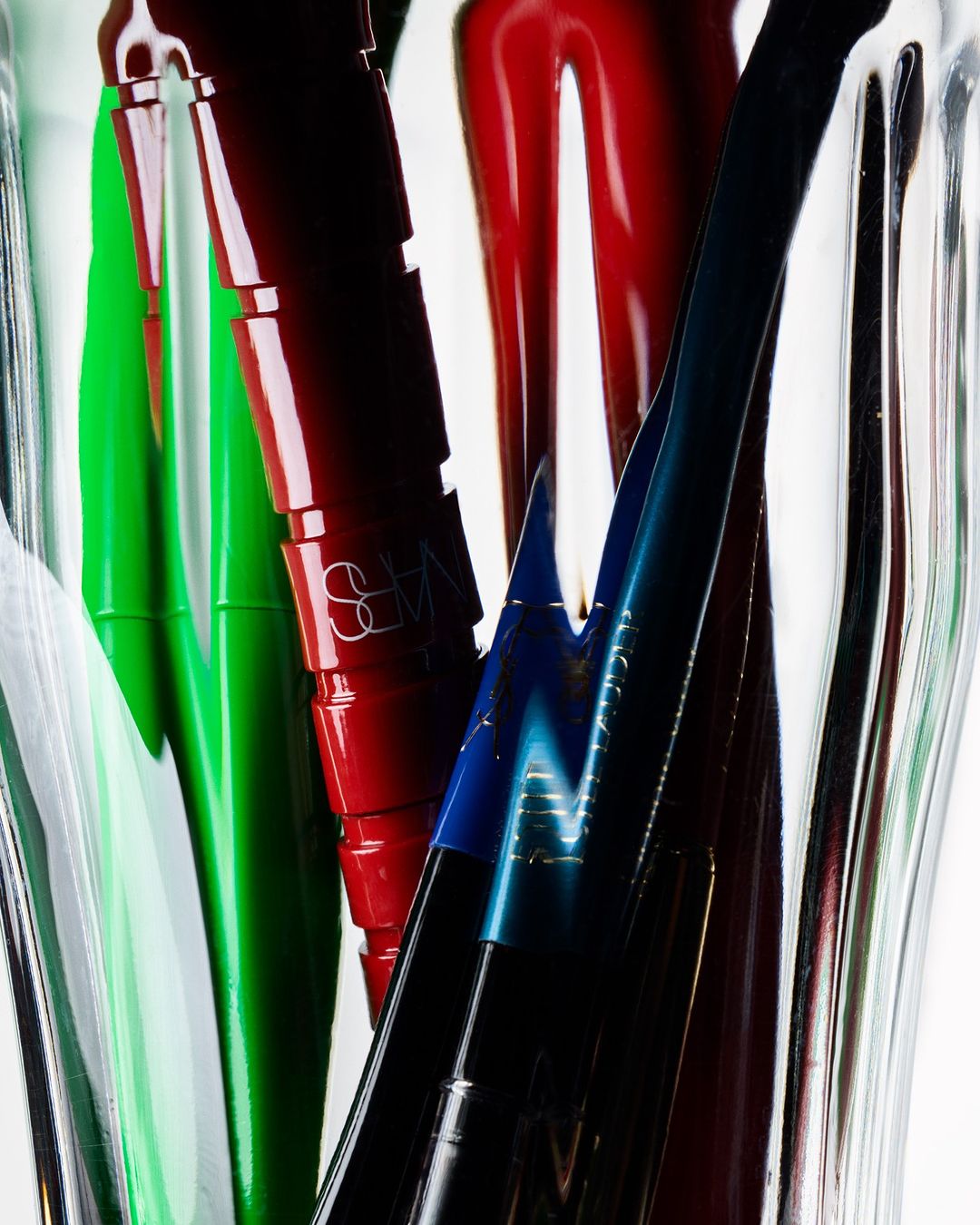 Macro shot of colorful NARS lipsticks in green, red, and blue shades in clear vase, against white background, for beauty advertising.