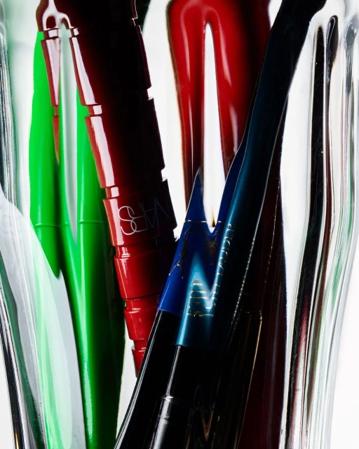 Macro shot of colorful NARS lipsticks in green, red, and blue shades in clear vase, against white background, for beauty advertising.