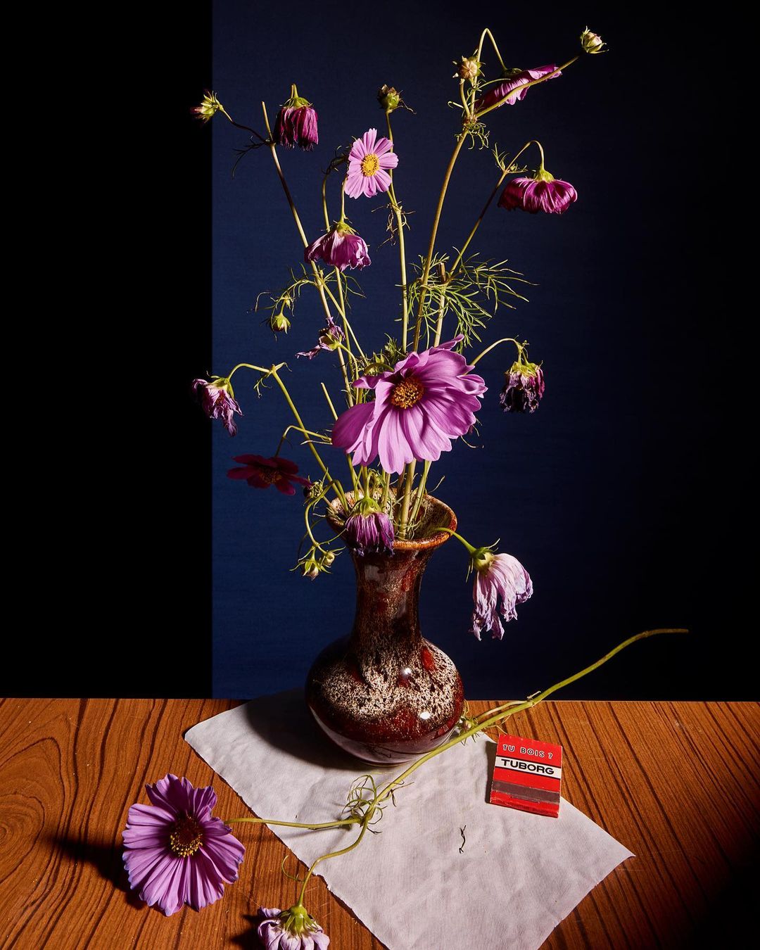 Still life of a flower arrangement in a vase on a wood table with a black and navy background, creating a somber and refined atmosphere.