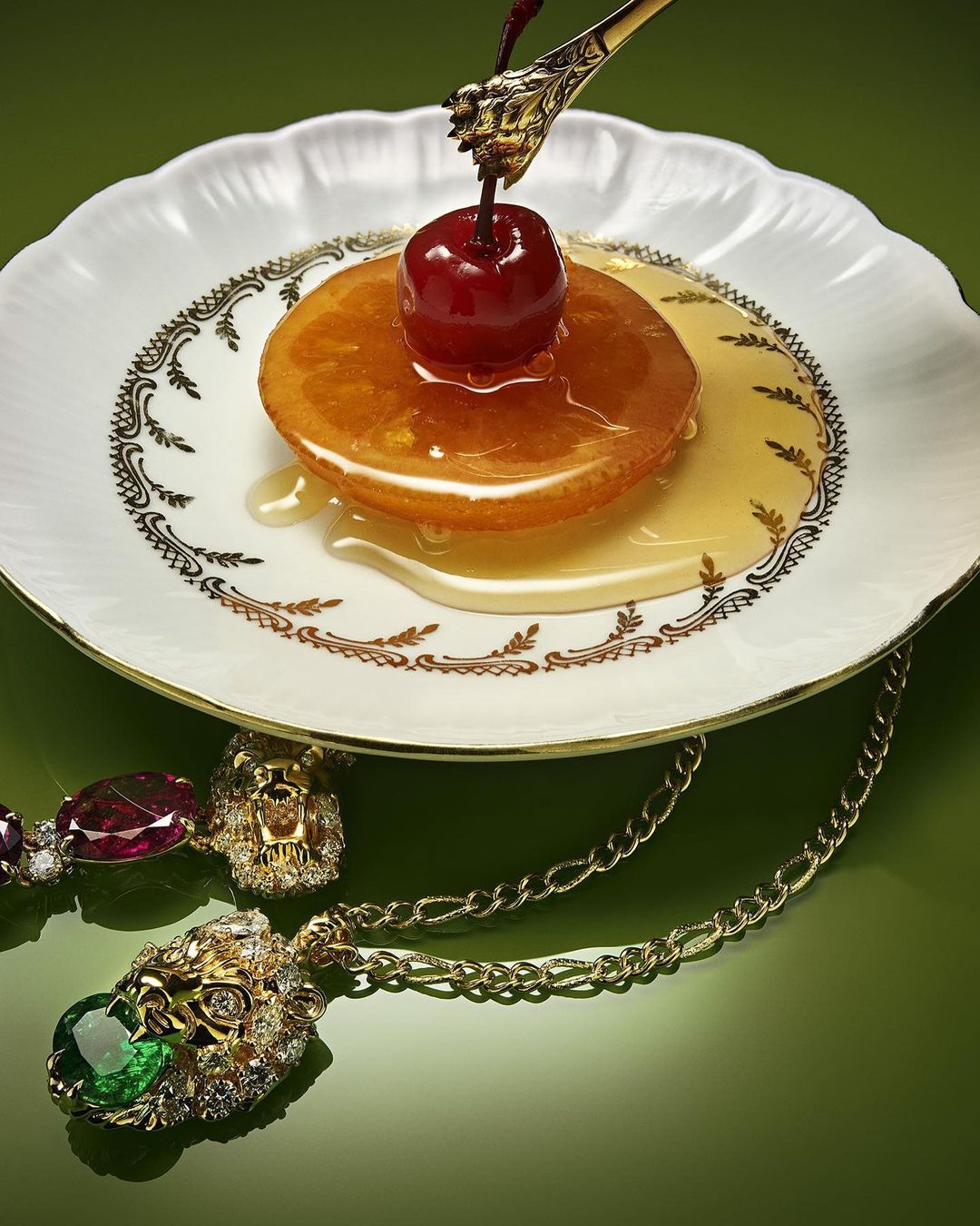 Overhead shot of a glazed fruit dessert with a cherry on a decorative plate, accented by a gold spoon and lion-themed necklace, on green.