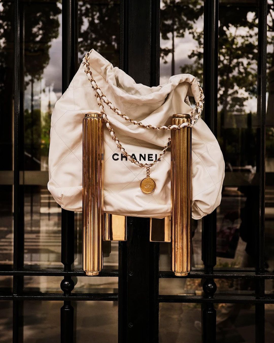 Chanel handbag and chain hanging on a gold metal door handle, photographed outdoors in natural light with trees reflected in the glass.