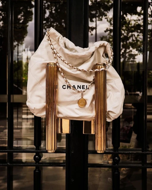 Chanel handbag and chain hanging on a gold metal door handle, photographed outdoors in natural light with trees reflected in the glass.