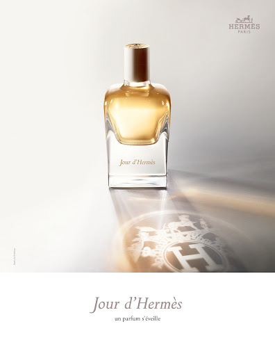 Elegant still life of a glass "Jour d'Hermès" perfume bottle on a shiny surface, reflecting the logo, against a gradient white background.