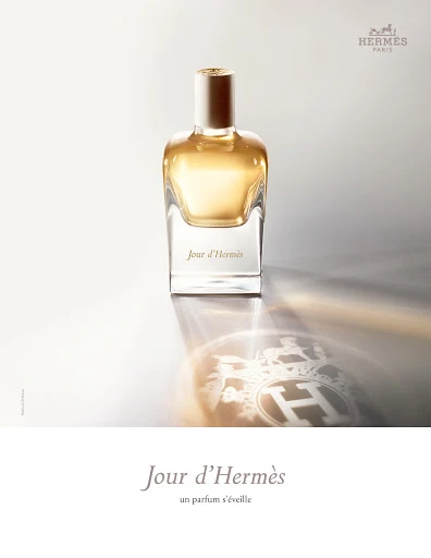 Elegant still life of a glass "Jour d'Hermès" perfume bottle on a shiny surface, reflecting the logo, against a gradient white background.