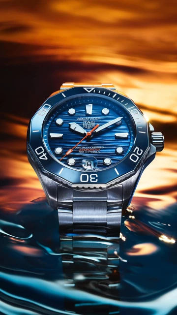 Close-up of a stainless steel TAG Heuer Aquaracer wristwatch with a blue dial, lying on a reflective surface with a warm background.