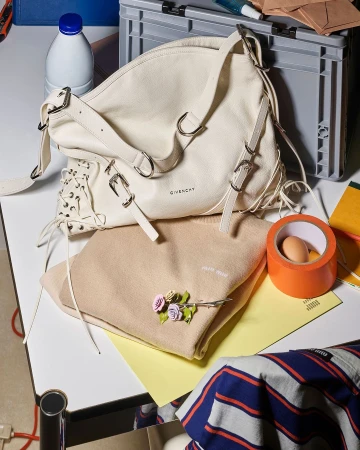 Cream-colored Givenchy handbag and tan Miu Miu sweater on a white table with an egg, tape, and blue-and-white striped shirt near a gray storage crate.