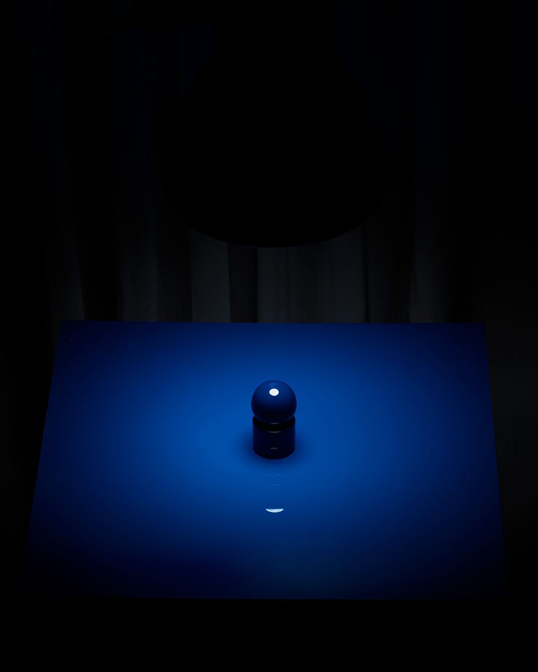 Still life photography of a blue sphere and cylinder object on a blue surface in front of a dark curtain background, studio lighting.