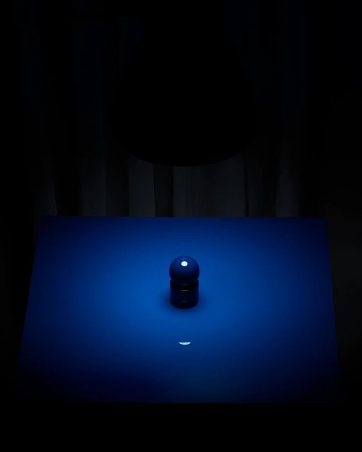 Still life photography of a blue sphere and cylinder object on a blue surface in front of a dark curtain background, studio lighting.