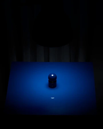 Still life photography of a blue sphere and cylinder object on a blue surface in front of a dark curtain background, studio lighting.