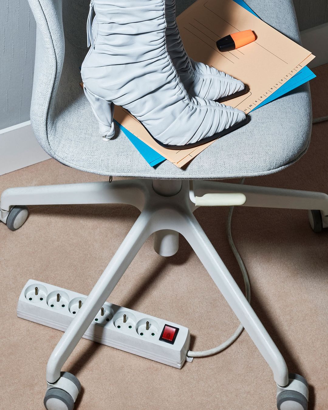 Office chair topped with gray high-heeled boots, folder, and highlighter, resting on a carpeted floor with a white power strip.