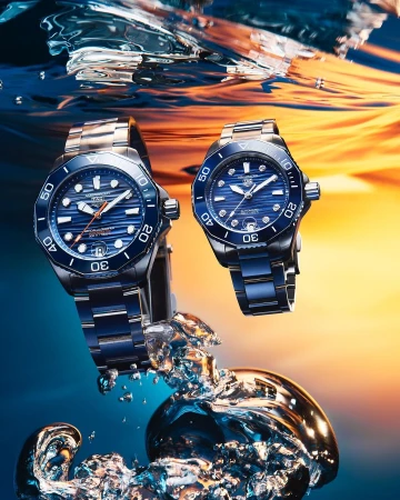 Two stainless steel TAG Heuer Aquaracer Professional 300 watches with blue dials, floating in water with a blue and orange background.