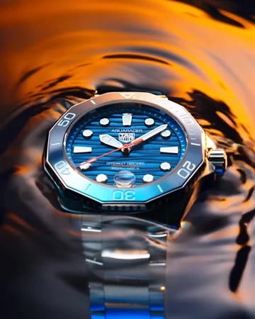 Close-up of a Tag Heuer Aquaracer wristwatch with a blue face and silver band partially submerged in water with an orange gradient background.