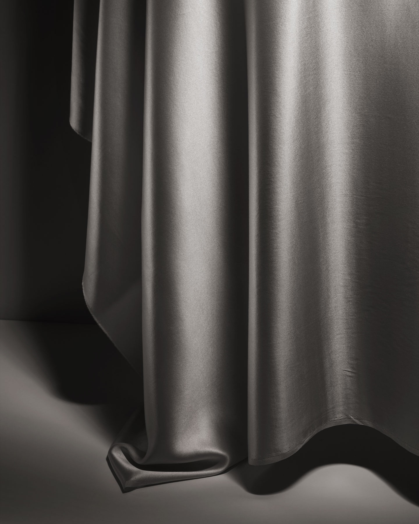 Abstract close-up of gray satin fabric draped on a gray surface, featuring soft lighting and shadows enhancing the material's sheen and texture.