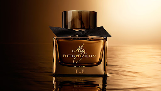 A luxurious amber-toned "My Burberry Black" perfume bottle with a black ribbon on a reflective surface against a softly lit gradient background.