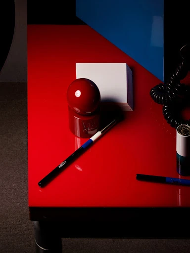 Still life photograph featuring a red table with a red sphere, white block, cosmetic brushes, and a black telephone cord.