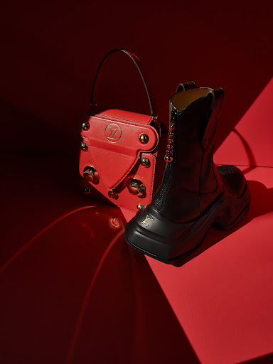 Elevated view: a red Louis Vuitton handbag with gold hardware next to a black leather combat boot on a glossy red surface.