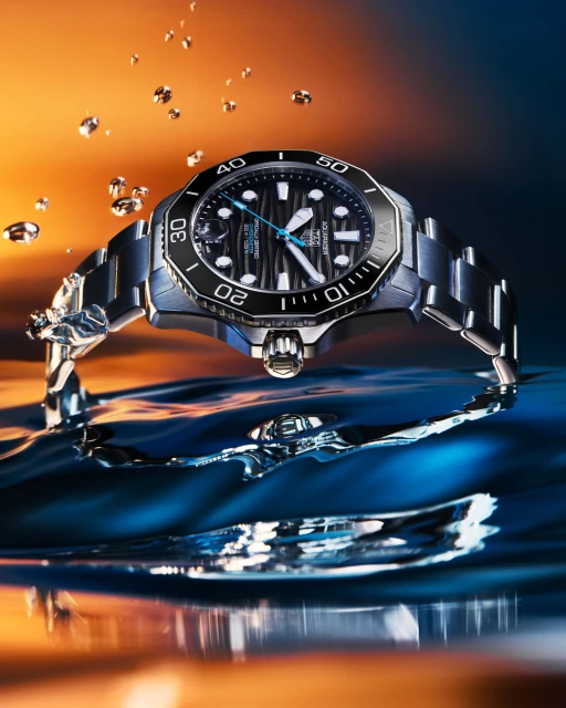 Elevated view of a silver wristwatch with a black face and bezel, reflecting in water, surrounded by droplets against an orange gradient background.