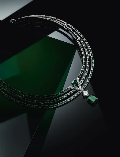Elegant silver necklace with emerald and diamond gemstones against a dark backdrop with subtle green reflective accents.