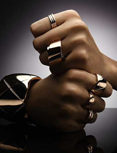 Close-up of two hands stacked, adorned with gold rings and a wide gold bangle against a blurred gray backdrop, emphasizing jewelry design.