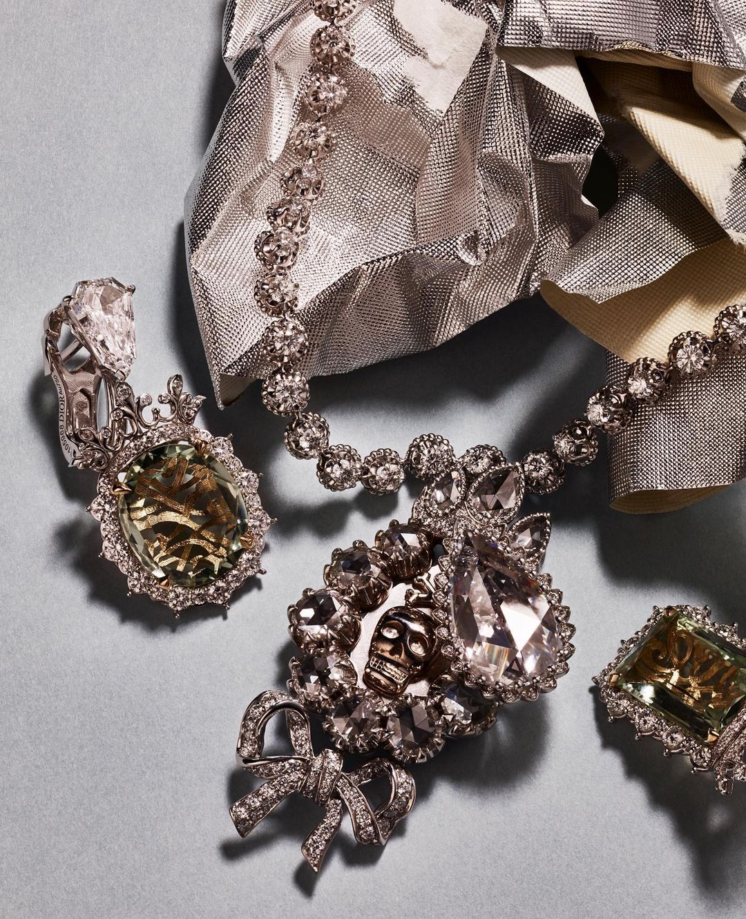 High-end jewelry: a diamond necklace, ring, and earring set on a light blue surface with silver crinkled wrapping in the background.