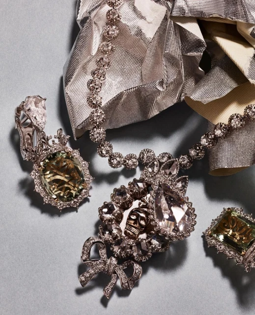 High-end jewelry: a diamond necklace, ring, and earring set on a light blue surface with silver crinkled wrapping in the background.