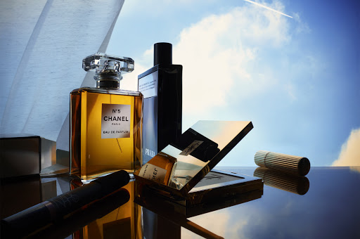 Still life shot of Chanel No. 5 perfume, Prada cologne, and Yves Saint Laurent makeup on a reflective surface with a sky background.
