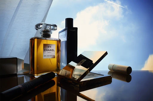 Still life shot of Chanel No. 5 perfume, Prada cologne, and Yves Saint Laurent makeup on a reflective surface with a sky background.