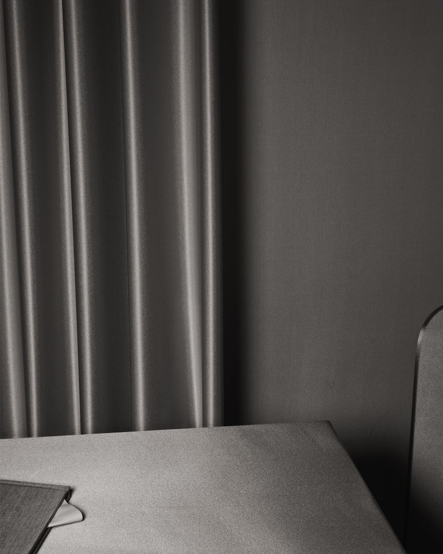 Minimalist interior of a room featuring gray textured drapes, a gray wall, a table edge, and the top of a gray chair.
