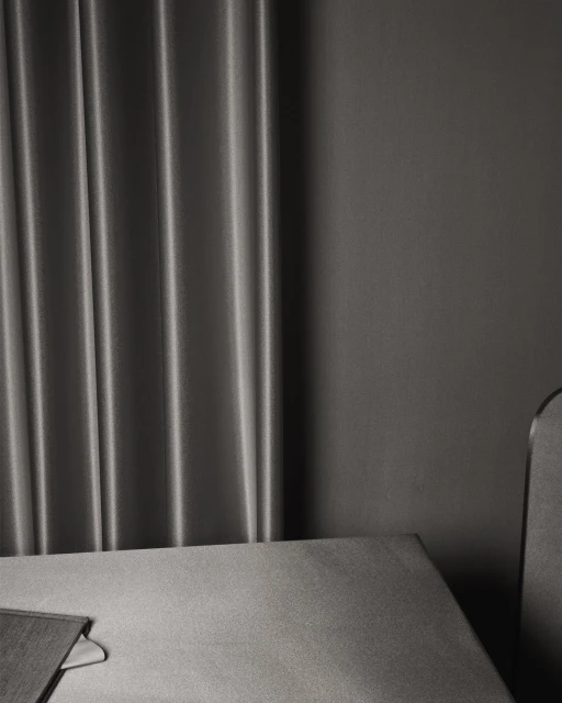 Minimalist interior of a room featuring gray textured drapes, a gray wall, a table edge, and the top of a gray chair.