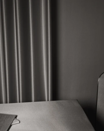 Minimalist interior of a room featuring gray textured drapes, a gray wall, a table edge, and the top of a gray chair.