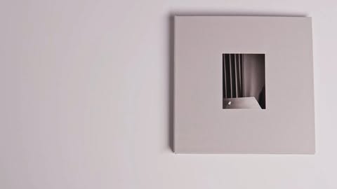Monochromatic photograph of a framed photo hanging on a wall, featuring an image of what appears to be a table against blinds.