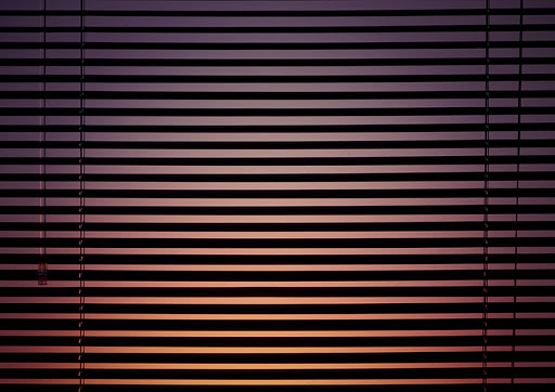 Closed window blinds filtering light into a room, creating horizontal lines and warm hues. Interior view during evening.