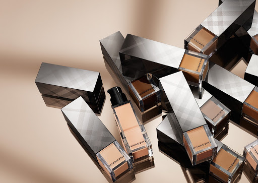 Burberry foundation makeup displayed on reflective surface with clear bottles, black pumps, and gray plaid packaging.