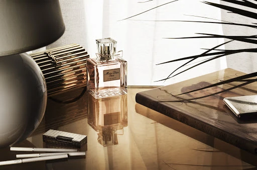 Still life shot of Miss Dior perfume bottle, cigarettes, lighter, striped clutch, and book, set on a reflective glass table.
