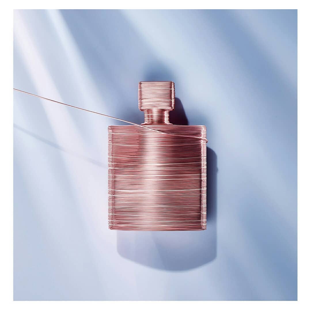 Artistic perfume bottle crafted from copper wire, set against a soft blue background with delicate light and shadow play, clean aesthetic.