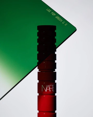Close-up product shot of a red Nars lipstick tube positioned vertically against a plain white background with a green filter.