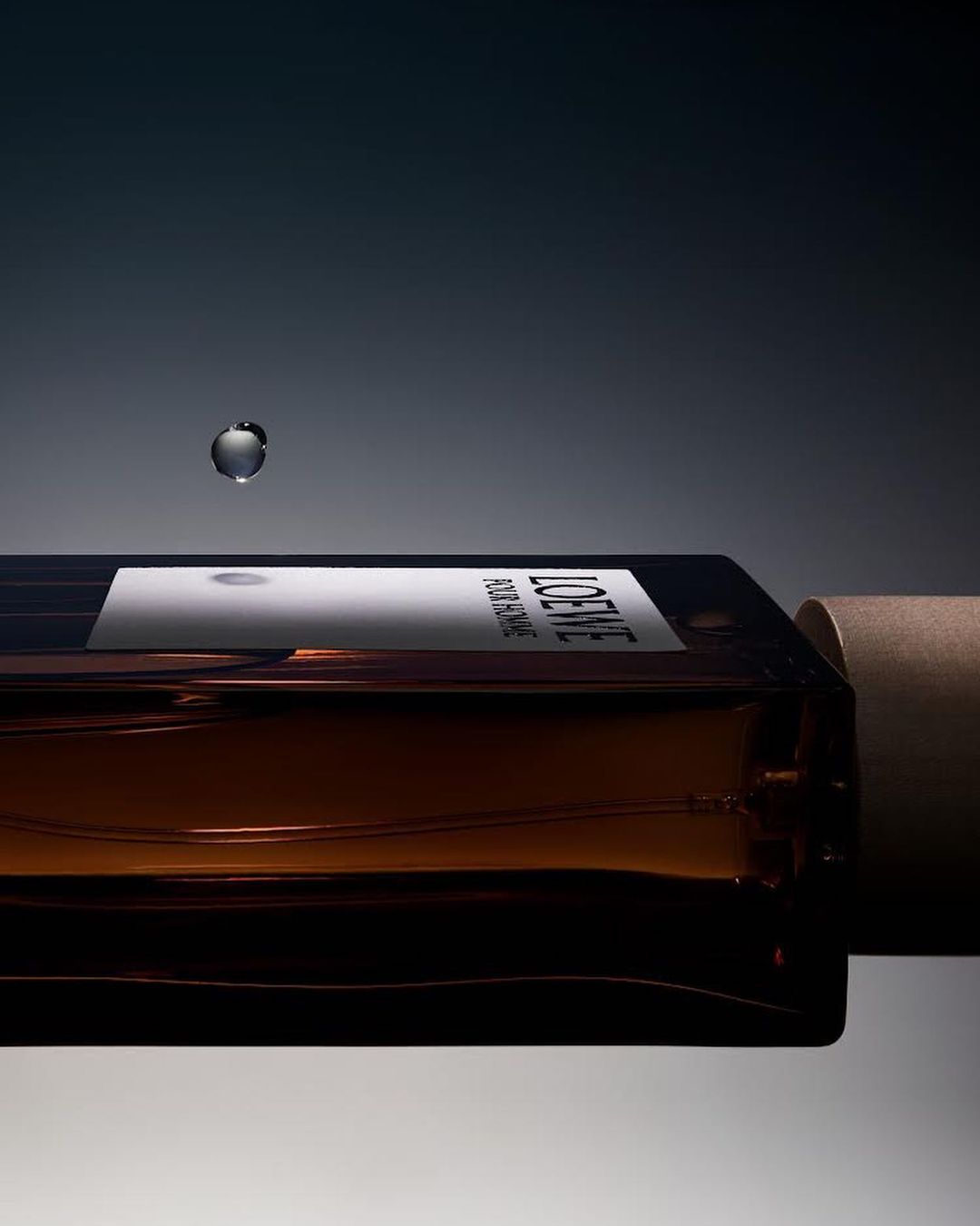 Loewe perfume bottle on its side with a water droplet hovering above it against a gradient grey and dark grey background.